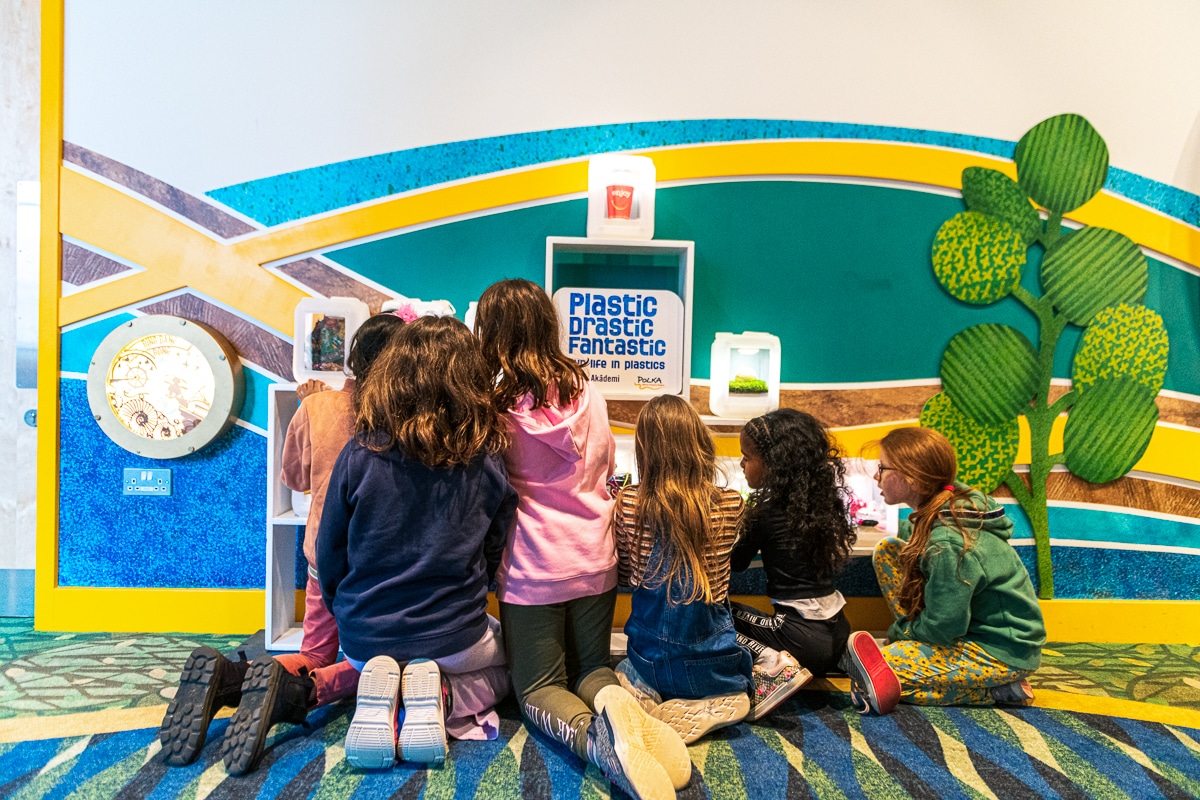 A group of children, kneeling in front of the Plastic Drastic Fantastic exhibition installation, gather together excitedly looking at the artwork with their backs to the viewer.